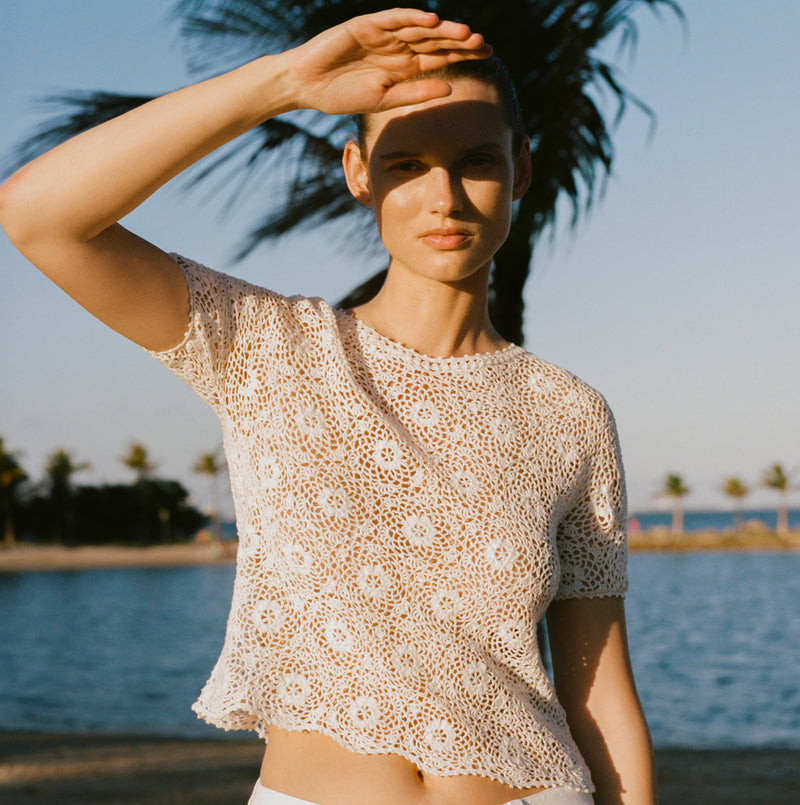 Woman in a lace top shielding her eyes from the sun by a palm tree.