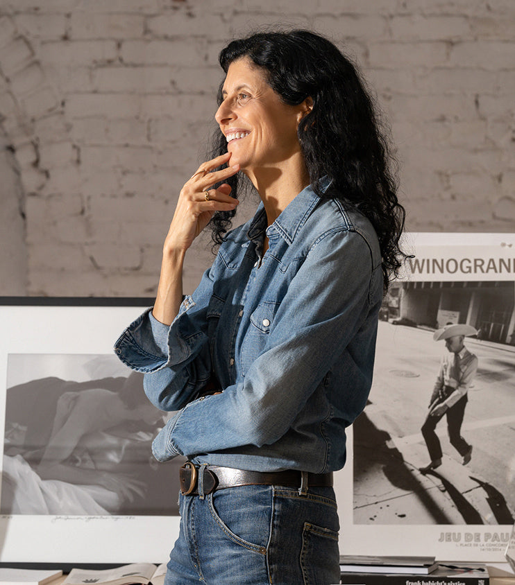 Woman smiling thoughtfully in a denim outfit, standing near framed photographs.