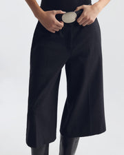 ILFORD GAUCHO WOOL PANT IN A close-up of a person wearing black wide-leg cropped pants with hands adjusting a belt, photographed against a light gray studio background.