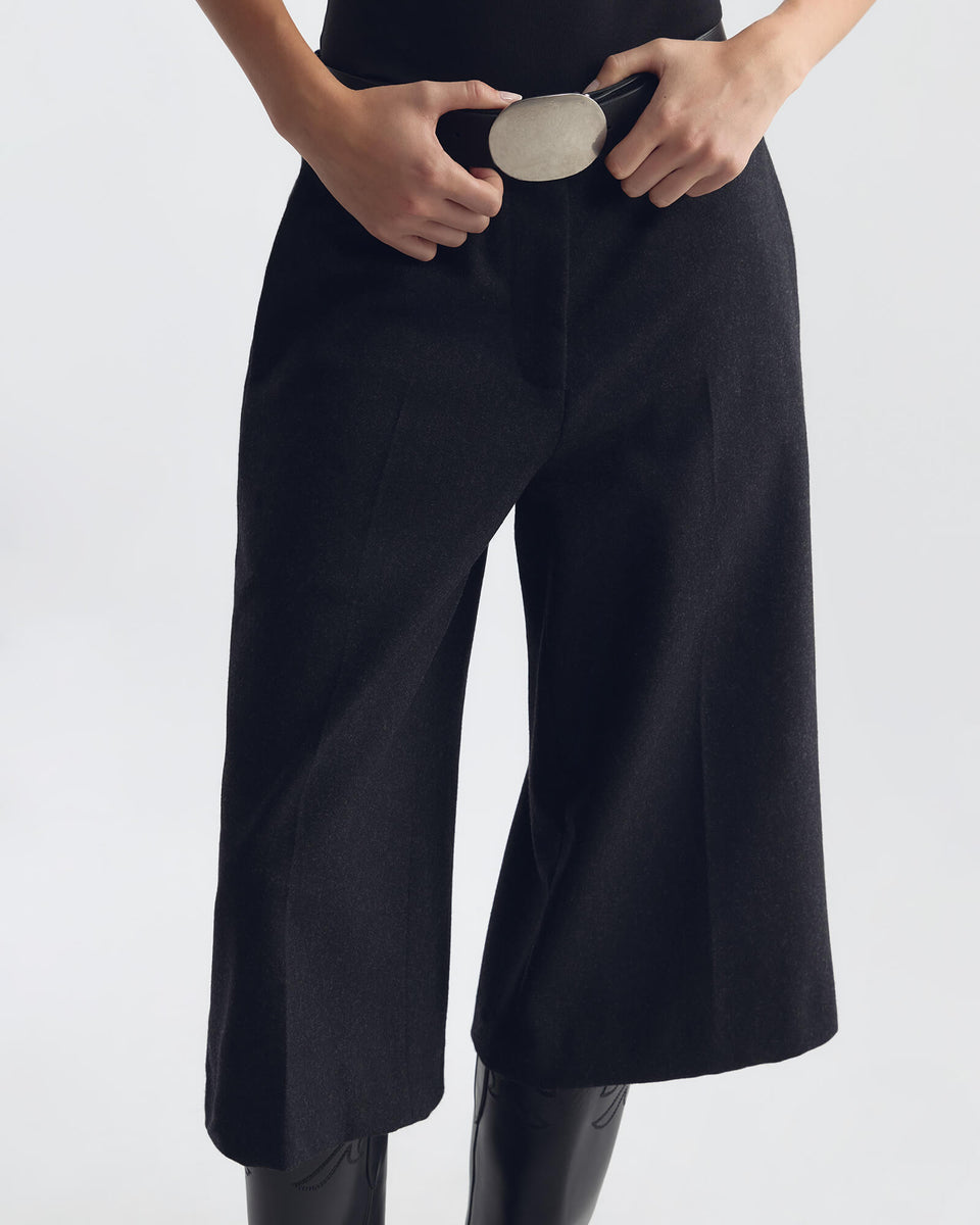 ILFORD GAUCHO WOOL PANT IN A close-up of a person wearing black wide-leg cropped pants with hands adjusting a belt, photographed against a light gray studio background.